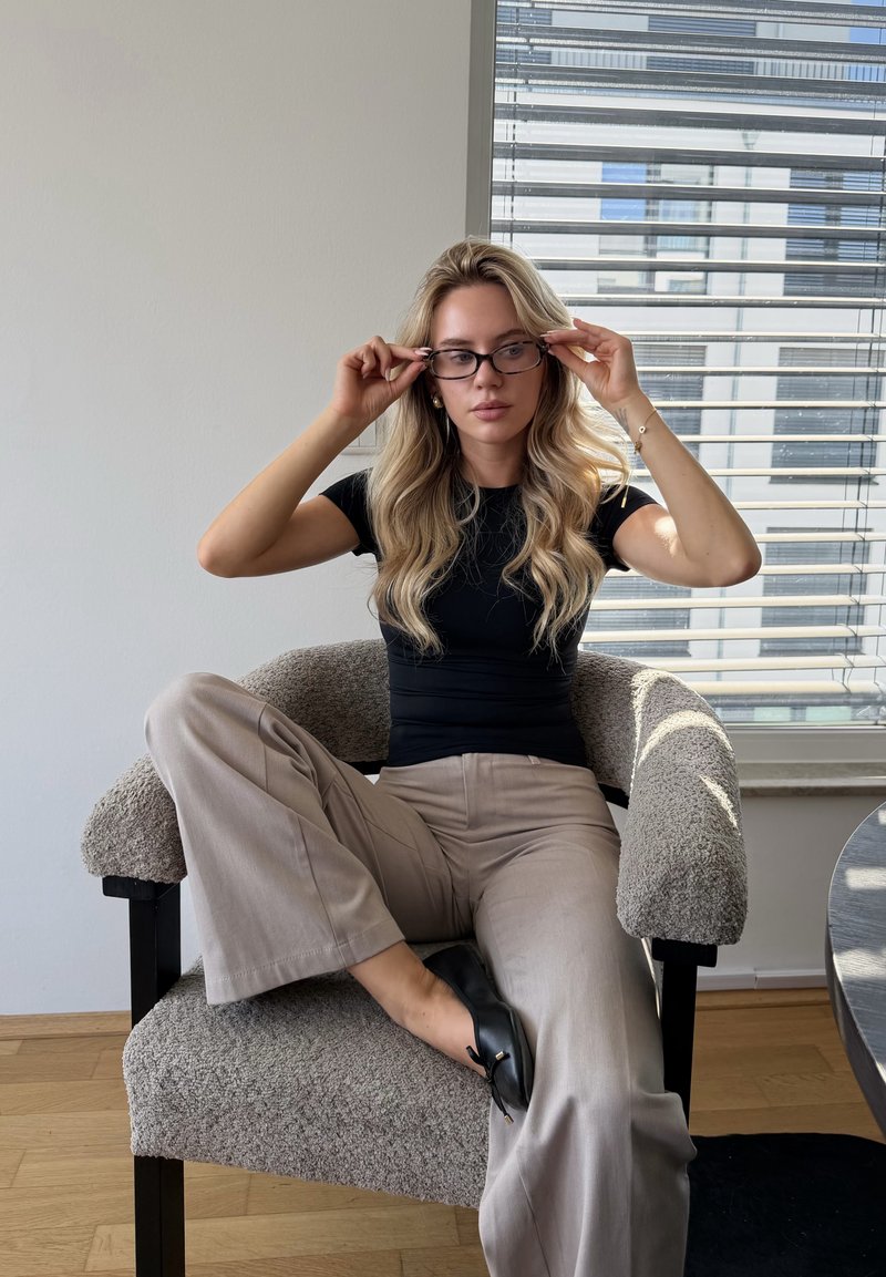 Femme aux longs cheveux blonds portant des lunettes, une chemise noire et un pantalon beige, assise en tailleur sur un fauteuil texturé près d'une fenêtre avec des stores.