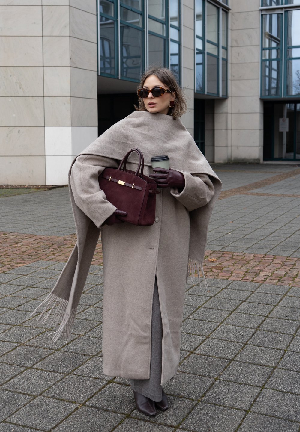 Manteau en laine beige à bords frangés, porté sur une tenue grise. Sac à main marron avec ferrures dorées, lunettes de soleil et tasse de café. Fond de trottoir gris.
