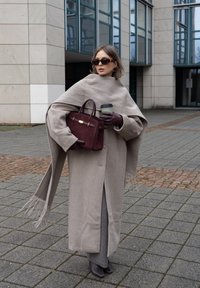 Manteau en laine beige à bords frangés, porté sur une tenue grise. Sac à main marron avec ferrures dorées, lunettes de soleil et tasse de café. Fond de trottoir gris.