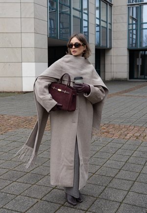 Abrigo de lana beige con bordes desflecados, llevado sobre un conjunto gris. Bolso marrón con herrajes dorados, gafas de sol y una taza de café. Fondo de pavimento gris.