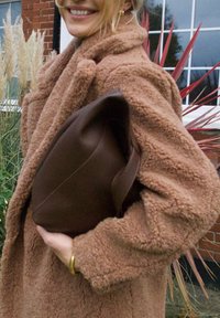 Brown leather handbag held against a fuzzy, tan coat with a shawl collar. The bag has a rounded shape and a short, sturdy handle.