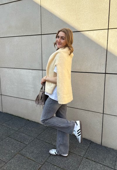 Capa de punto amarillo claro con bordes acanalados, llevada sobre una camisa blanca, combinada con jeans de campana grises y sneakers en blanco y negro.