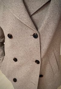 Kétsoros, világosszürke gyapjúkeverékből készült kabát fekete gombokkal, enyhén kivágott gallérral és oldalzsebekkel, texturált szövésű anyagból.