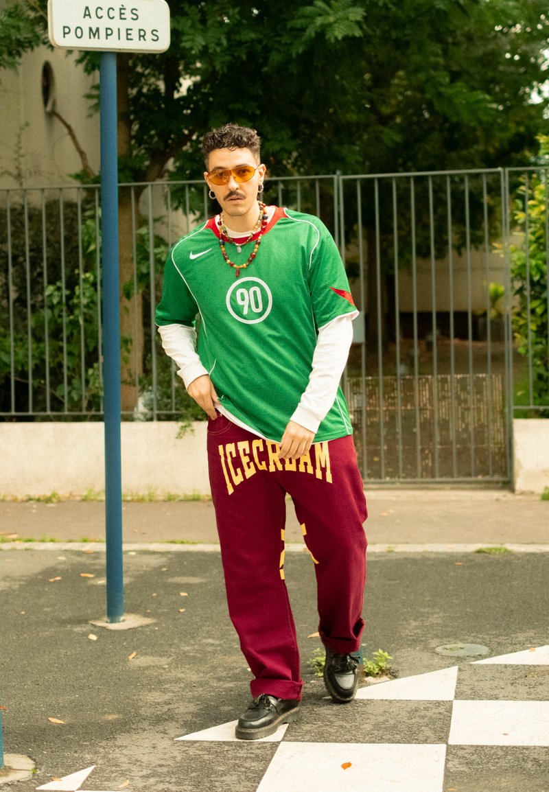 Camiseta verde de mangas curtas com um logotipo circular, combinada com calças de treino bordô com letras amarelas. Usada com sapatos pretos e acessórios.
