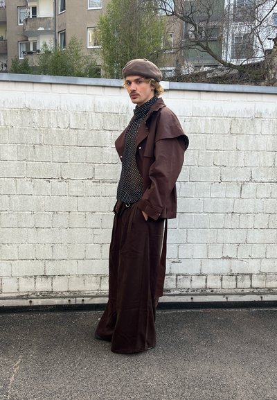 Joven de pie junto a una pared blanca de ladrillos, vistiendo una boina marrón, una chaqueta marrón de gran tamaño, pantalones de pierna ancha y una camisa negra con lunares.