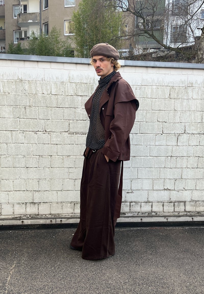 Młoda osoba stoi przy białej ceglanej ścianie, ubrana w brązową beretkę, brązową oversize'ową kurtkę, spodnie o szerokich nogawkach i czarną koszulę w groszki.