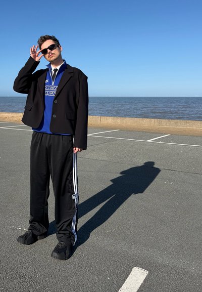 Hombre con gafas de sol, blazer negro, camisa azul y pantalones deportivos negros Adidas de pie sobre la acera junto al mar bajo un cielo despejado.