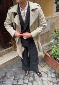 Bež kabanica preko tamnog odijela s prugicama, upotpunjena smeškim kožnim cipelama. Figura prilagođava dugme na kabanici.