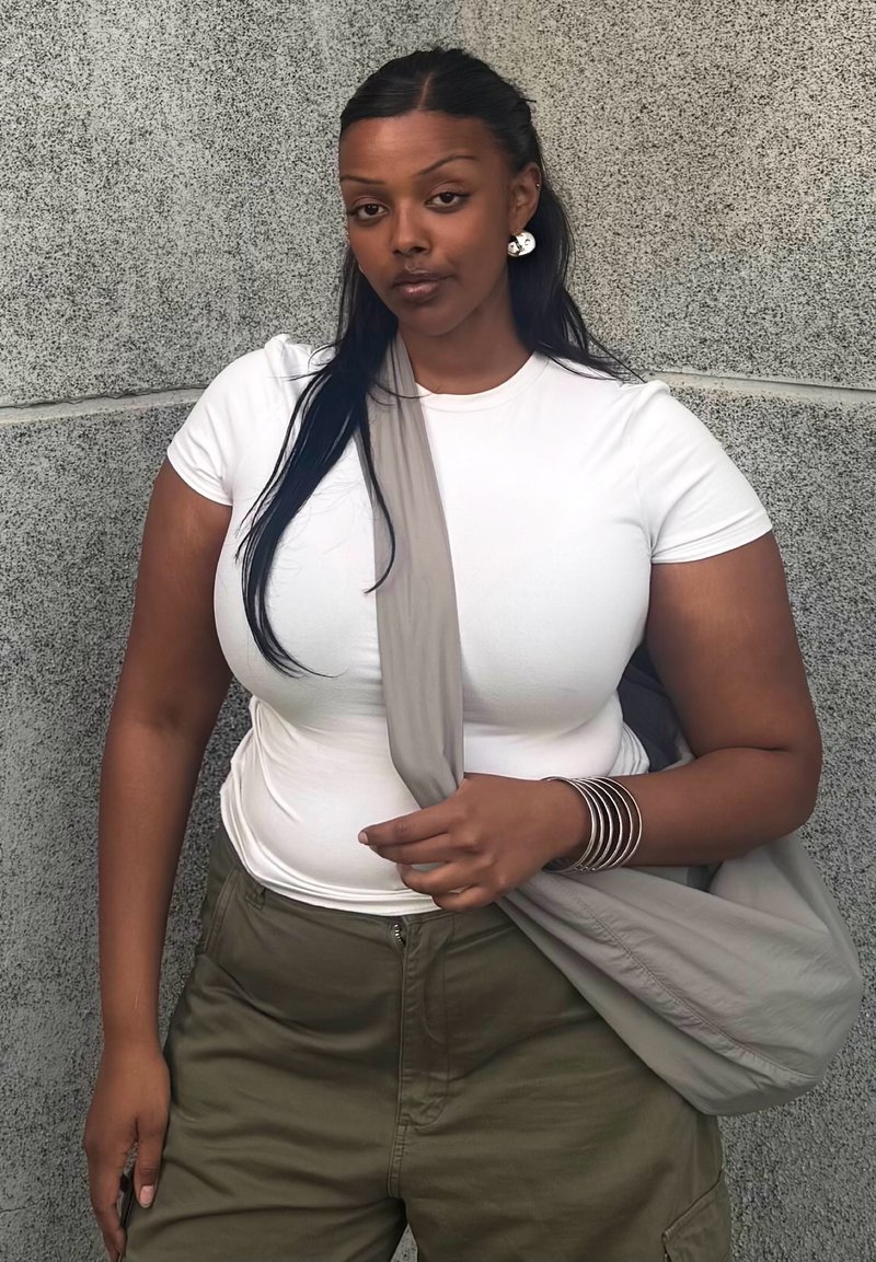 Femme aux longs cheveux noirs portant un t-shirt blanc, un pantalon olive, des boucles d'oreilles argentées, des bracelets et un sac gris porté en bandoulière, debout contre un mur gris.