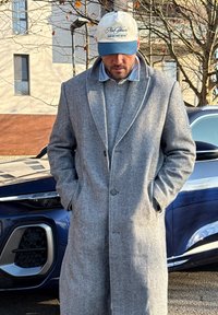 Homme portant un pardessus en chevrons gris et une casquette blanche et bleue, debout les mains dans les poches à côté d'une voiture bleu foncé en milieu urbain.