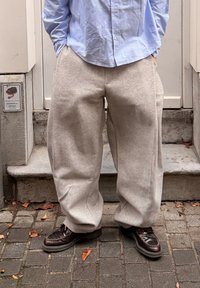 Šviesiai pilkos spalvos, plačiomis kelnėmis, pagamintos iš minkštos, tekstūruotos medžiagos. Derinamos su tamsiai rudos spalvos batais, stovinčios ant akmenimis grįsto paviršiaus.