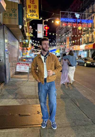 Chaqueta de lona marrón sobre una camisa blanca, pantalones cargo azules y zapatillas de deporte navy. Entorno urbano con coloridos letreros de neón y luces de cuerda.