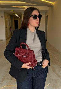 Czarny żakiet w paski, szara bluzka, ciemne dżinsy; trzyma torebkę w burgundowym kolorze o teksturowej powierzchni z zamkiem; nosi czarne okulary przeciwsłoneczne i czerwone kolczyki.