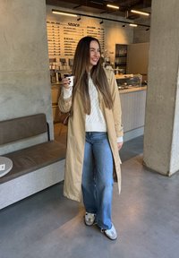 Mulher sorridente com cabelo longo castanho, vestindo um trench coat bege, suéter branco e jeans, segurando uma chávena de café dentro de um café moderno.