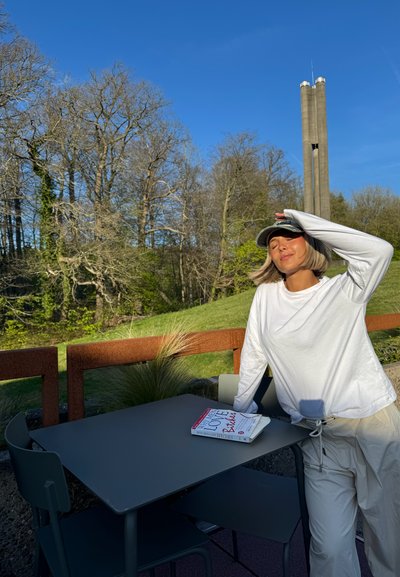 Camisa blanca de manga larga, pantalones claros y gorra; mesa gris con un libro titulado "Amor perras". El fondo presenta árboles y una alta estructura de concreto.