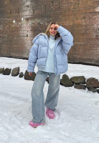 Femme souriante en doudoune bleu clair et jean, debout sur la neige près d’un mur en pierre avec un graffiti à la craie. Bottines roses visibles.