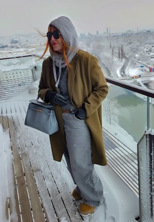Mujer con abrigo de invierno, sudadera con capucha, guantes y gafas de sol, de pie en un balcón nevado con vistas al río y al paisaje urbano en invierno.