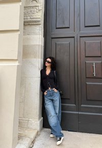 Kvinne i svart knappet topp, vide blå jeans og joggesko lener seg mot steinvegg ved stor mørk tredør, iført solbriller og tverrveske.