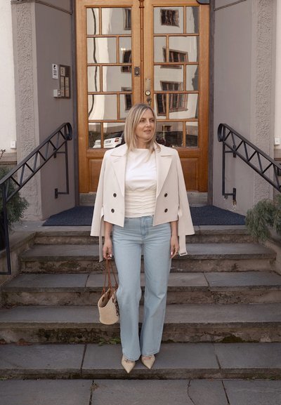 Blazer entallado de color claro con doble botonadura sobre una blusa blanca, combinado con jeans acampanados azul claro y un bolso beige tejido.