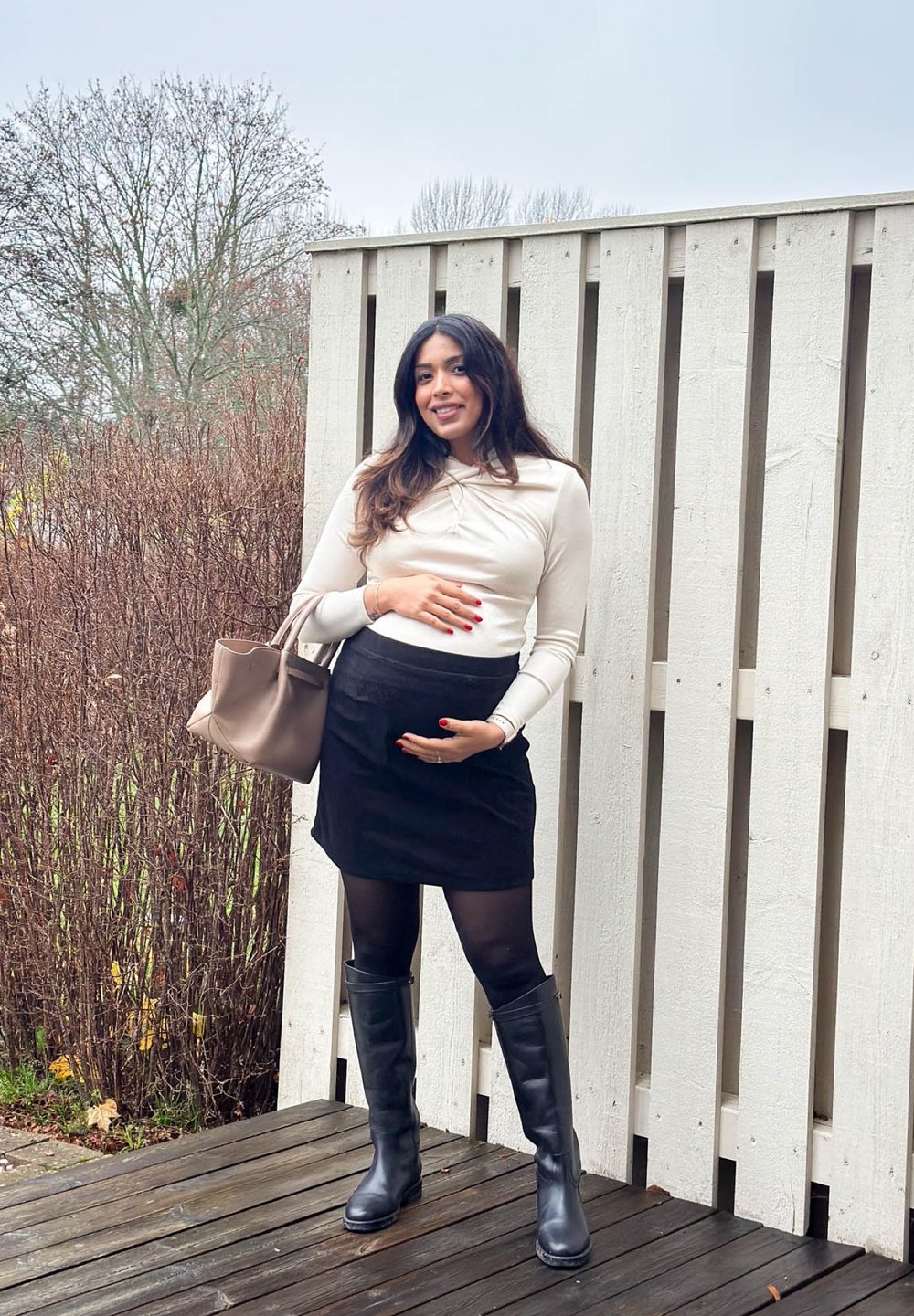 Lichtbeige top met lange mouwen, zwarte minirok, zwarte ondoorzichtige panty's en kniehoge zwarte laarzen. Draagt een beige handtas, staande in de buitenlucht.