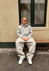 Harmaa ylijättöhuppari ja sitä vastaavat collegehousut, jotka on valmistettu pehmeästä materiaalista. Valkoiset lenkkarit. Henkilö istuu puisella penkillä teksturoidun seinän edessä.