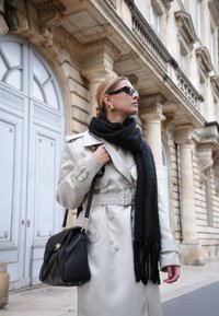 Beige Trenchcoat mit Gürtel, schwarzer Schal und Sonnenbrille. Schwarze Handtasche. Der Hintergrund zeigt klassische Architektur mit Säulen.