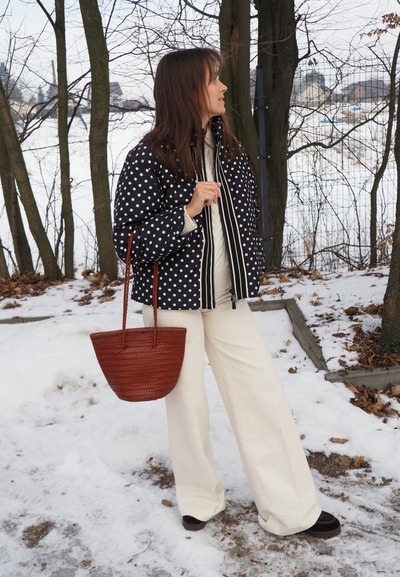 Femme portant une veste noire à pois et un pantalon crème, tenant un sac tressé marron, debout sur un sol enneigé près des arbres.