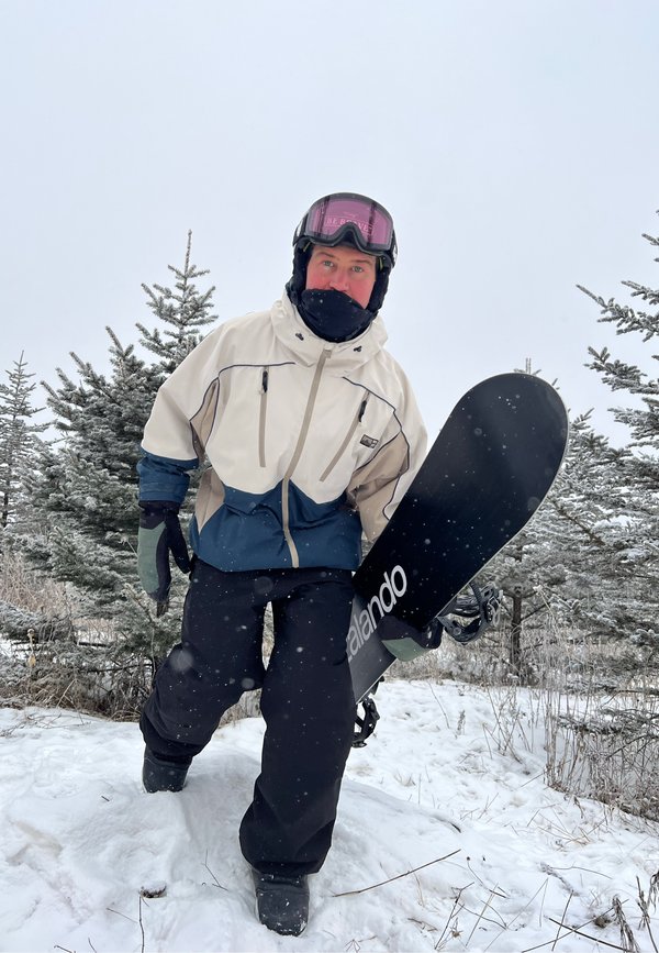 Personne en tenue d'hiver tenant un snowboard, debout sur un sol enneigé avec des pins couverts de neige en arrière-plan.