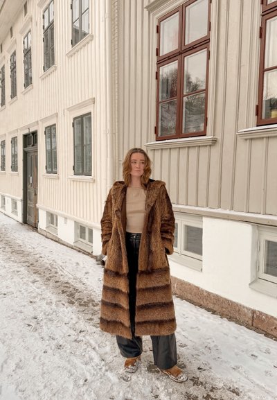 Mujer de pie en una calle nevada, vistiendo un abrigo largo de piel marrón, pantalones negros y botas color canela, al lado de un edificio beige con varias ventanas.