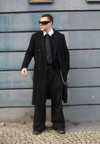 Long manteau noir sur un costume noir avec cravate, orné d'un détail chaîne. Lunettes de soleil et bague argentée complètent la tenue devant un mur bleu.