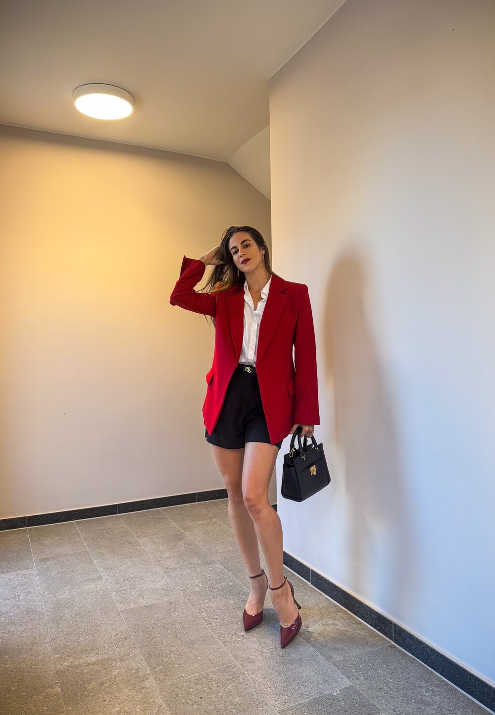 Rød blazer over en hvit skjorte, kombinert med svarte shorts. Tilbehør inkluderer en svart håndveske og burgunder høyhælte sko. Glatt, nøytral bakgrunn.