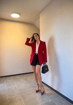 Blazer rojo sobre una camisa blanca, combinado con pantalones cortos negros. Los accesorios incluyen un bolso negro y tacones burdeos. Fondo suave y neutro.