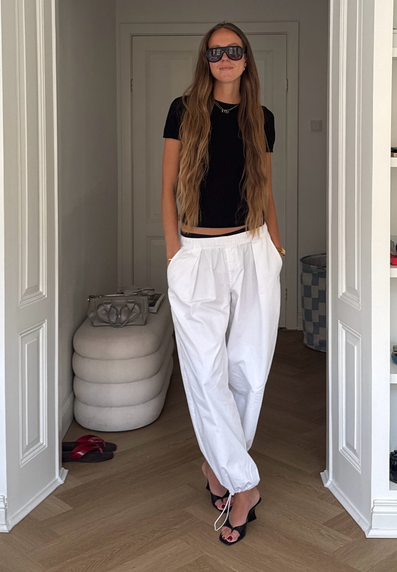Kvinne med langt hår iført svart topp, hvite bukser, svarte sandaler med hæl og store solbriller, står i en døråpning med hendene i lommene.