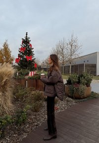 Brązowa pikowana kurtka, czarne welurowe spodnie, trzyma filiżankę. W tle choinka z czerwonymi kokardami oraz donice z zielonymi roślinami.