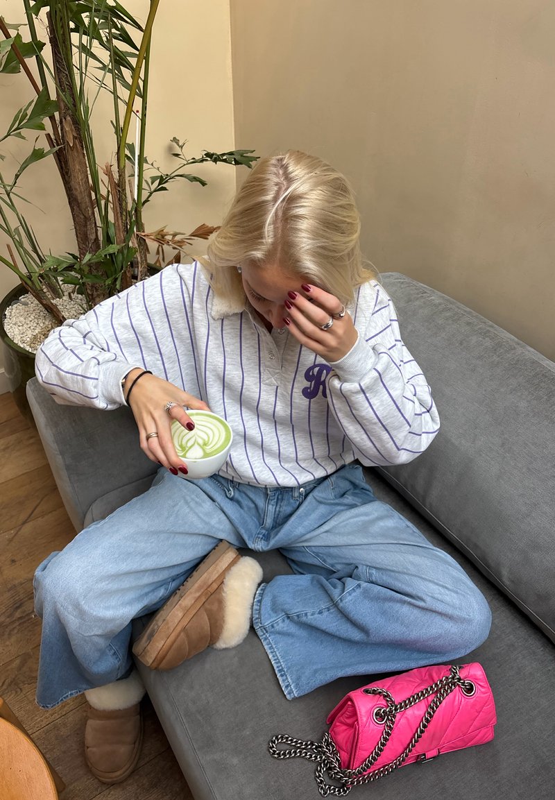 Chemise à manches longues rayée blanc et violet, jean large bleu clair, chaussures pantoufles couleur fauve, tenant une boisson tourbillonnante verte dans une tasse blanche, sac à main rose.
