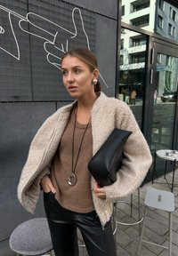 Chaqueta de teddy beige y esponjosa con mangas anchas, combinada con un suéter de punto marrón y pantalones de cuero negros. Bolso de mano negro y detalle de colgante circular.