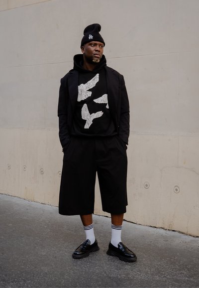 Sudadera negra de gran tamaño con gráfico de pájaro blanco, blazer negro, pantalones cortos holgados, zapatos negros y calcetines a rayas blancas. Fondo oscuro y pared neutra.