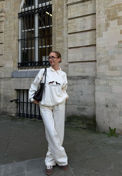 Sudadera blanca con cuello y diseño gráfico, combinada con pantalones anchos blancos. Zapatillas marrones y un bolso negro texturizado.