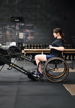 Mujer en silla de ruedas usando una máquina de remo en un gimnasio con pesas y equipo de ejercicio en el fondo.