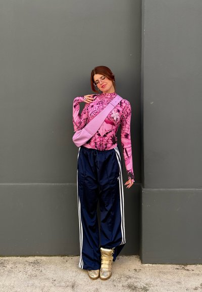 Top rosa de manga larga con patrones gráficos negros, pantalones de chándal azul marino con rayas blancas y botas metálicas doradas. Bolso cruzado rosa.