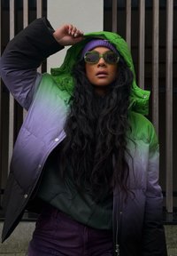 Σακάκι πούφ φτιαγμένο με γκράντ από πράσινο σε μοβ, με κουκούλα, ελαστικά μανίκια και γυαλιστερή επιφάνεια. Φορεμένο με πράσινα γυαλιά ηλίου και μοβ σκουφάκι.