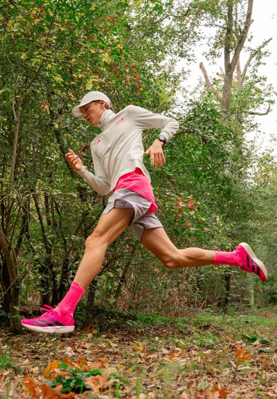 Corredor masculino con una camiseta de manga larga gris claro y pantalones cortos rosas, usando calcetines y zapatillas rosas, corriendo a través de un área boscosa.