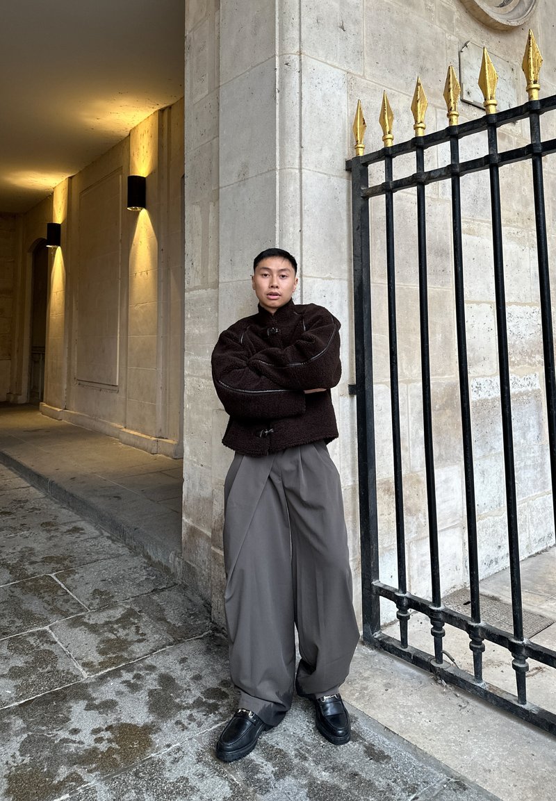 Pulover maro cu textură, supradimensionat, pantaloni largi gri și pantofi negri masivi. Stă în picioare lângă un zid de piatră cu o poartă neagră cu vârfuri aurii.