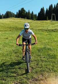 Széles gumikkal rendelkező mountain bike fekete vázzal és zöld kiegészítőkkel. A kerékpáros fehér sisakot és szürke pólót visel, zöld, füves kültéri környezetben.