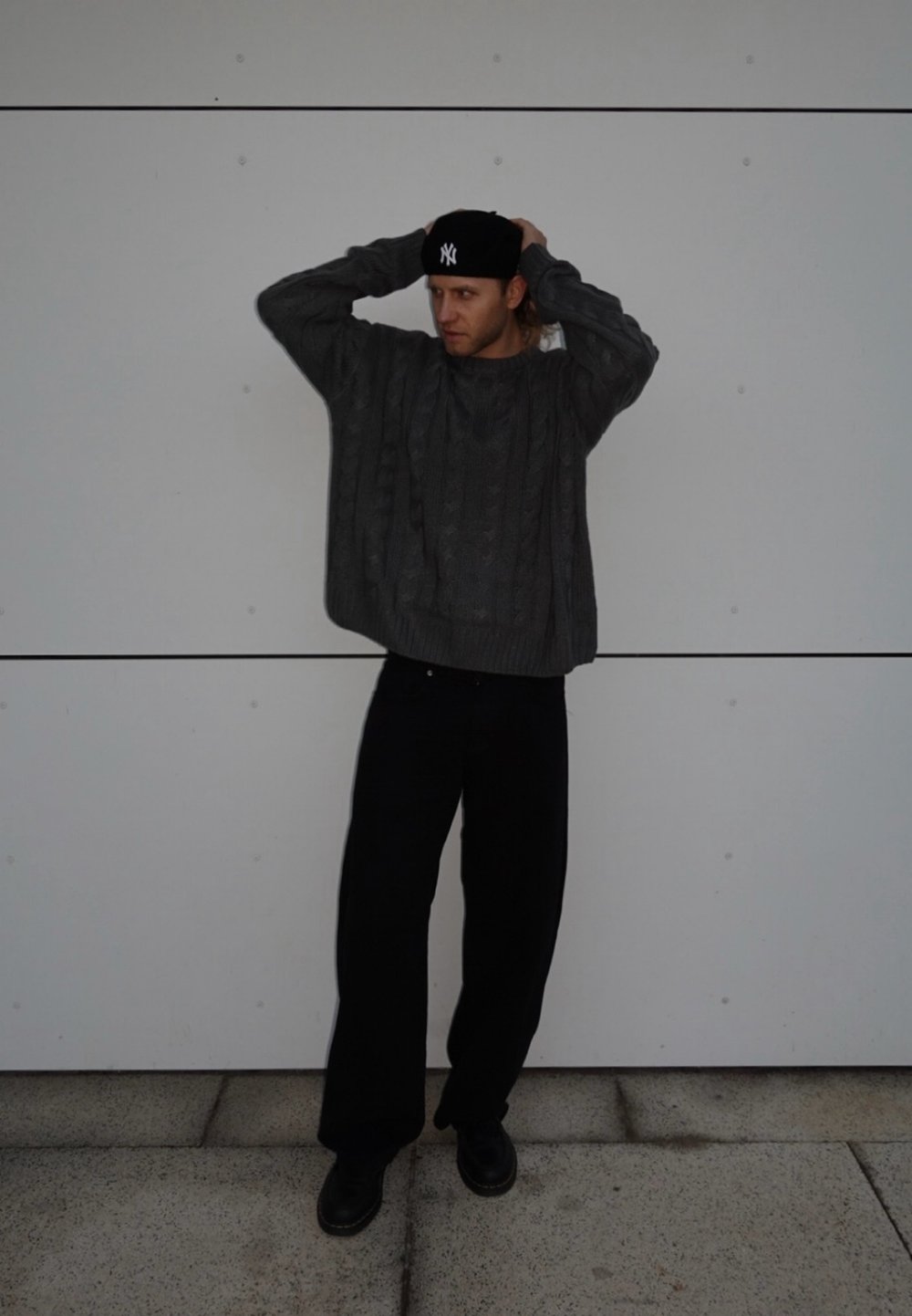 Grauer Kabelstrickpullover, schwarze lockere Hose und schwarze Schuhe. Die Person trägt eine schwarze Mütze, hat die Arme erhoben vor einem schlichten weißen Hintergrund.