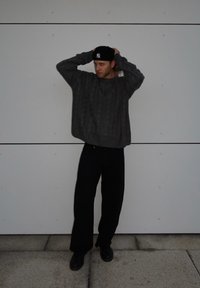 Grauer Kabelstrickpullover, schwarze lockere Hose und schwarze Schuhe. Die Person trägt eine schwarze Mütze, hat die Arme erhoben vor einem schlichten weißen Hintergrund.