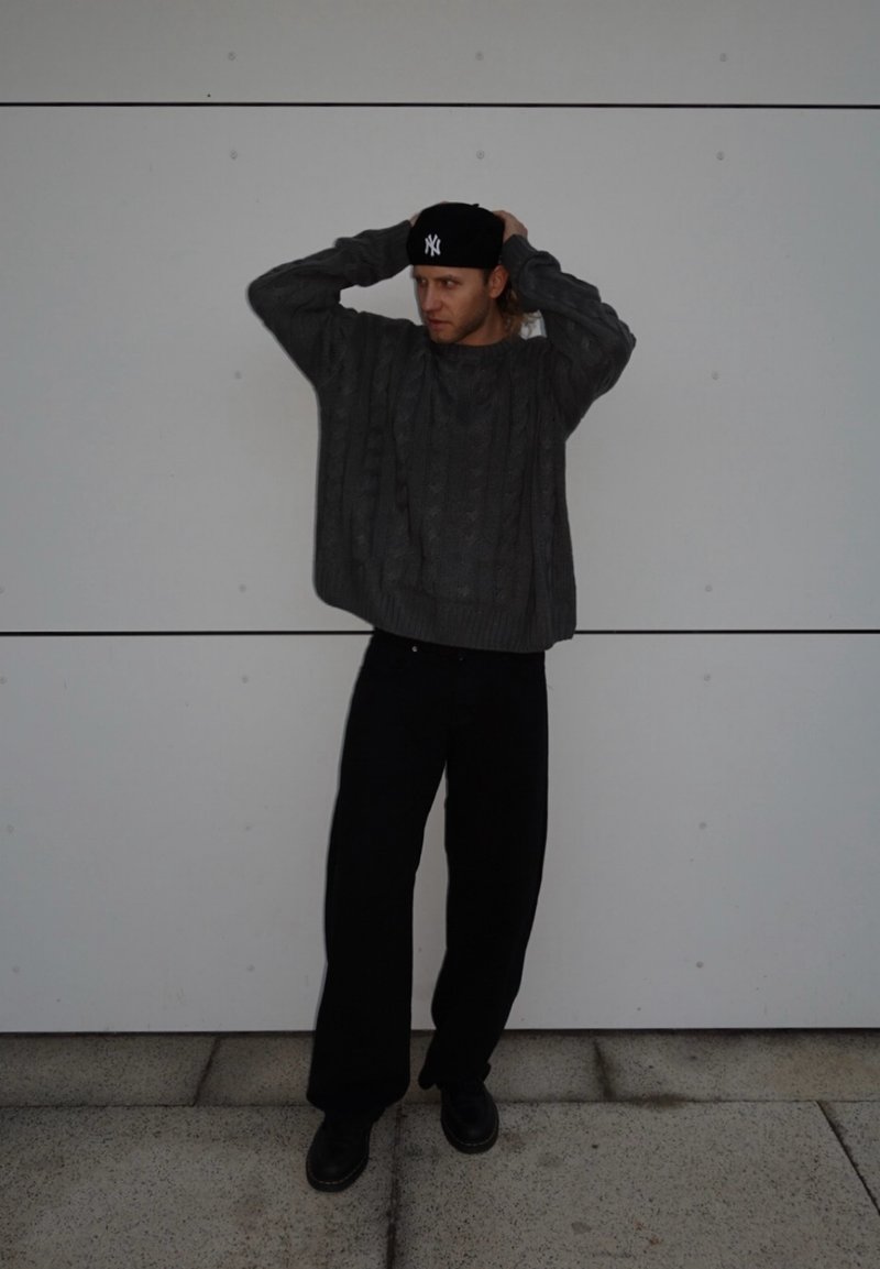 Grauer Kabelstrickpullover, schwarze lockere Hose und schwarze Schuhe. Die Person trägt eine schwarze Mütze, hat die Arme erhoben vor einem schlichten weißen Hintergrund.