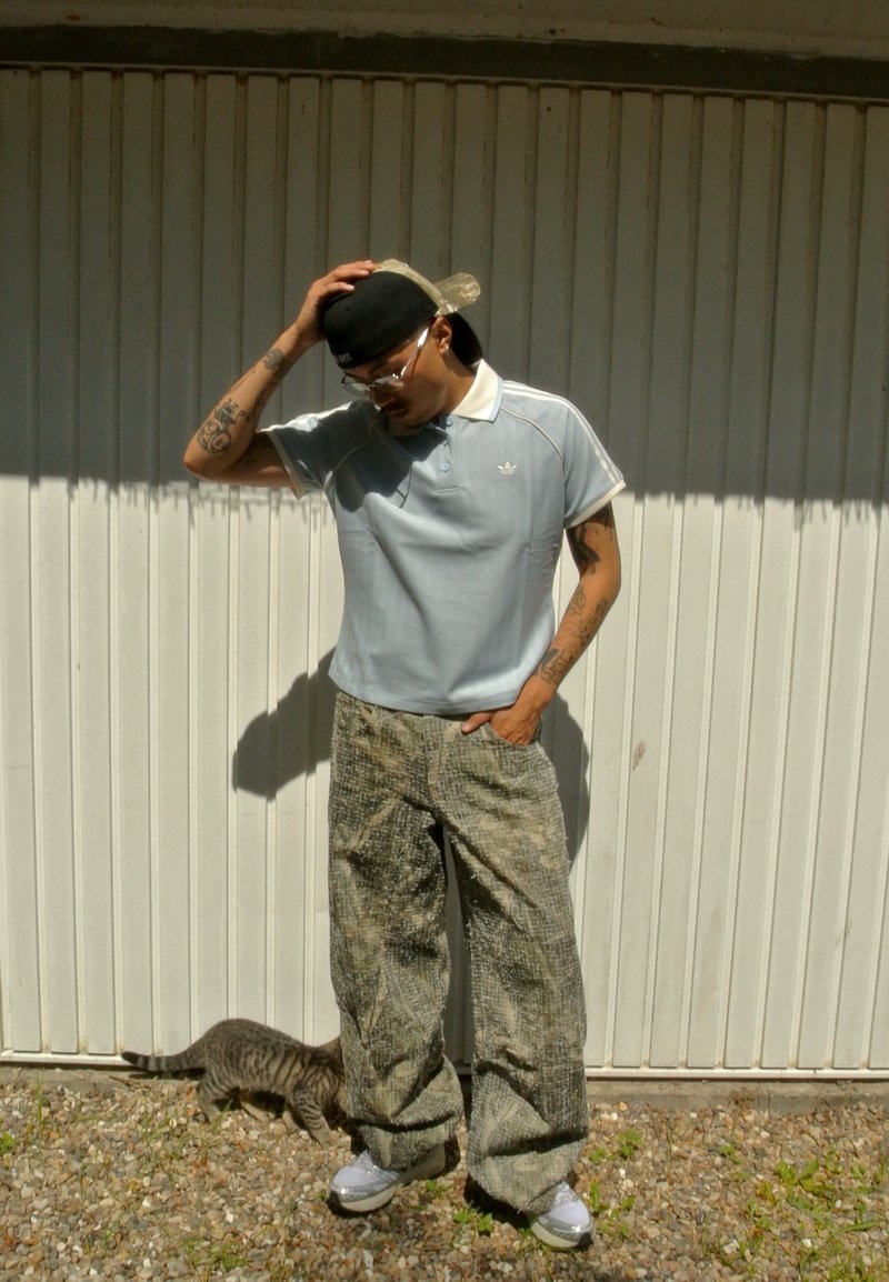 Homme tatoué portant une casquette, des lunettes, un polo bleu clair et un pantalon ample, debout contre un mur blanc avec un chat tigré qui renifle près de ses pieds.