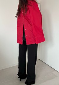 Röd överdimensionerad skjorta med knäppning, med en vänster bröstficka, matchad med svarta vida byxor och svarta klackskor, mot en ljus bakgrund.