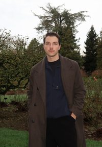 Manteau en laine brun porté par-dessus une veste zippée marine, associé à un pantalon noir. Scène située dans un jardin avec des arbres et de la verdure.